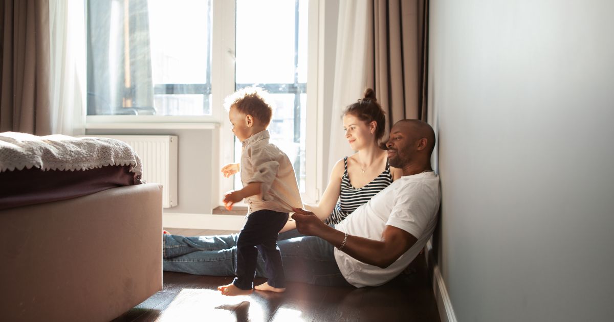 Couple sitting on the floor on their bedroom while toddler walks around.