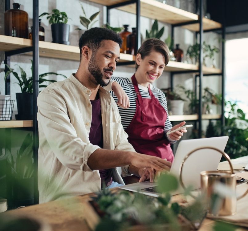 Woman and man business owners viewing phone and laptop in a flower shop.