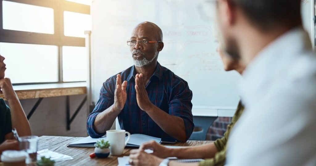 Man with glasses sitting in front of a whiteboard in a meeting with colleagues.